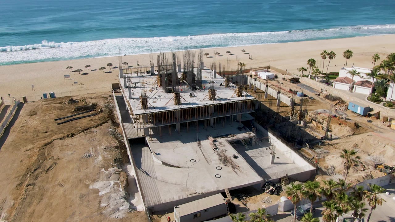 At 0:01, the El Tule Beach Los Cabos frame centers the rebar-filled structure, with umbrellas on the sand, palms to the right, ocean waves behind, and no visible people.