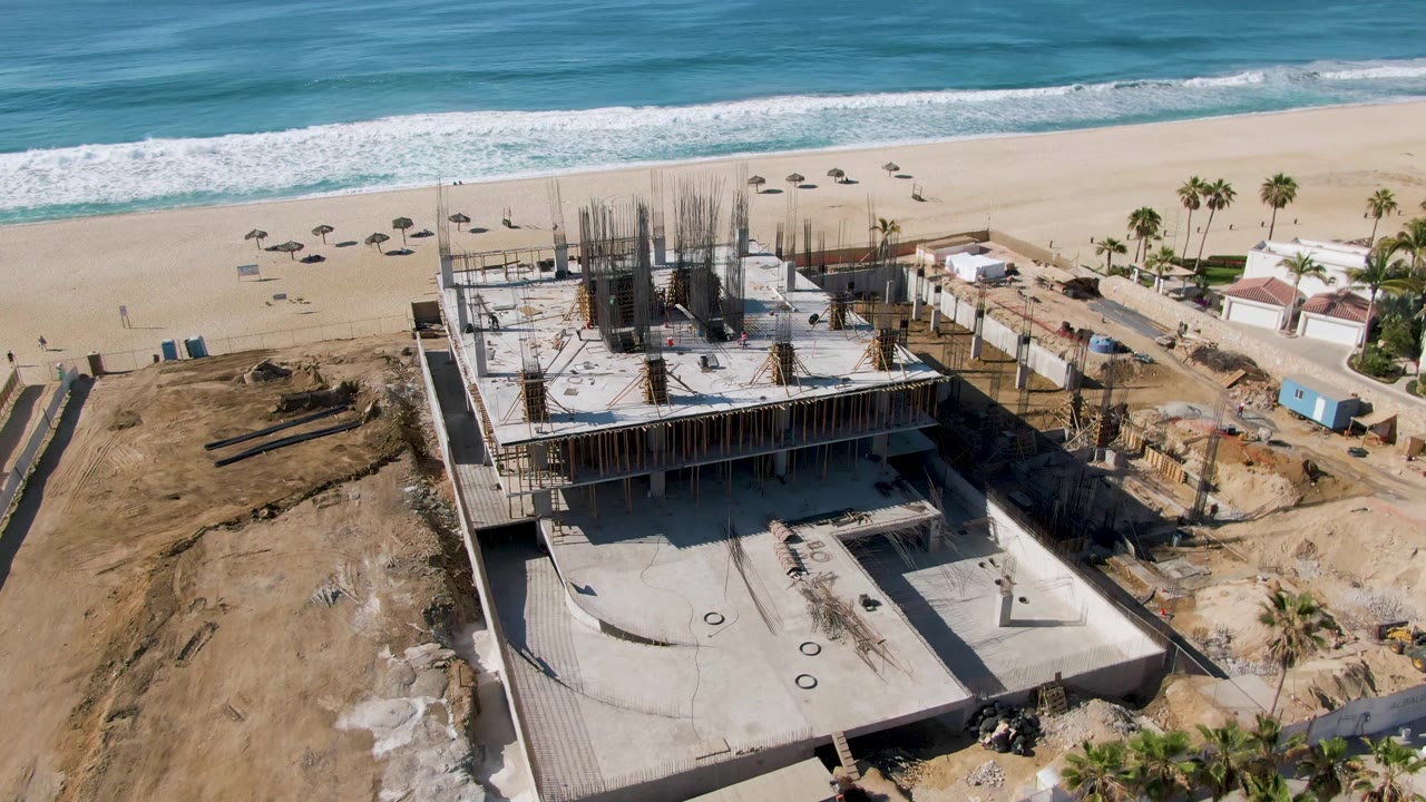 At 0:02, the drone view at El Tule Beach Los Cabos keeps the construction site in the lower frame while broad sand, white surf, palm trees, and no visible people fill the background.