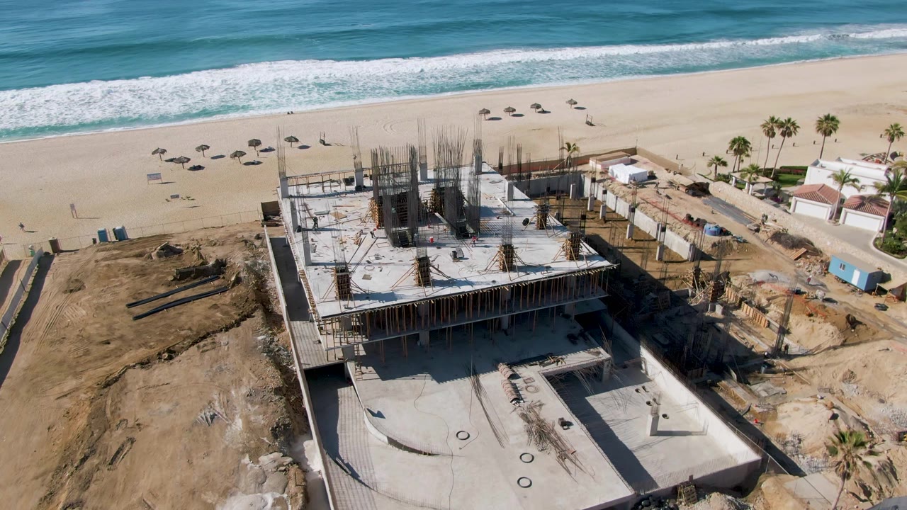 At 0:03, El Tule Beach Los Cabos appears from above with the unfinished concrete structure lower in frame, a wide strip of sand, breaking waves, palm trees, and no visible people.