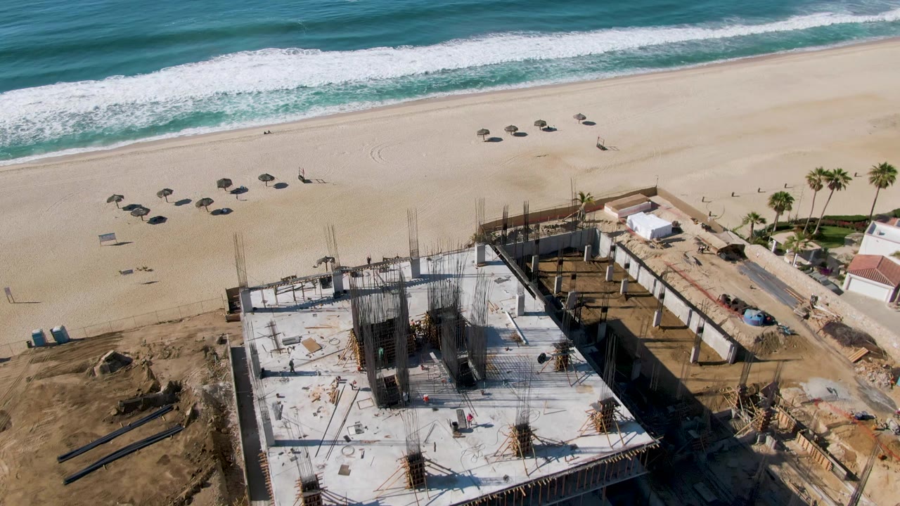 Aerial frame over El Tule Beach in Los Cabos showing teal surf, pale sand, scattered umbrellas, a few distant people, and a concrete construction site with rebar in the lower foreground.