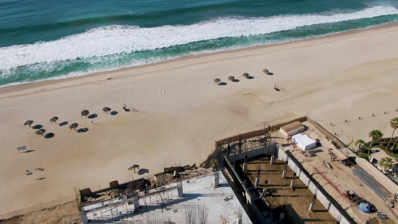 Aerial frame over El Tule Beach where the ocean and sand dominate, with umbrella clusters on the beach and the construction site reduced to the lower edge of the view.