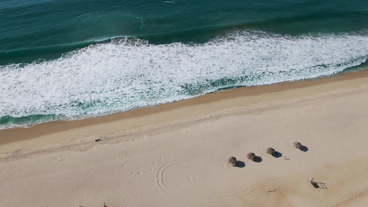 Overhead view at El Tule Beach in Los Cabos shows green-blue surf breaking toward pale sand, with small dark rocks or beach objects visible and no visible people.