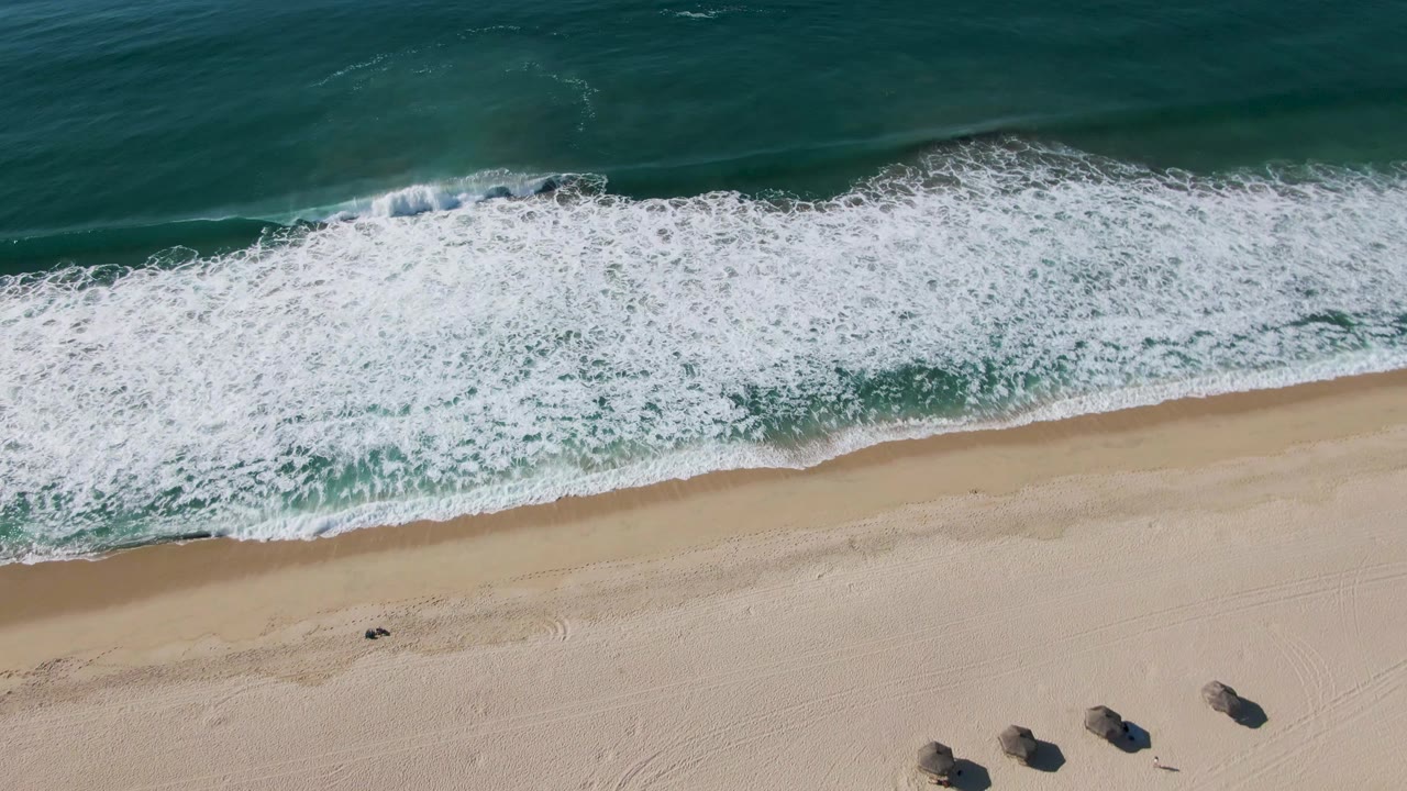 Aerial view at El Tule Beach in Los Cabos shows a wider white foam line moving across the sand, with dark beach objects near the lower right and no visible people.