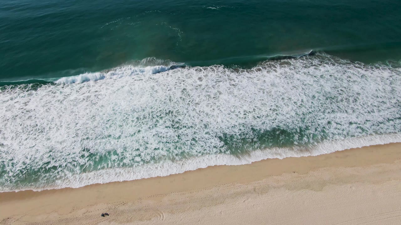 At El Tule Beach in Los Cabos, dense white surf fills most of the shoreline from above while green ocean water remains visible beyond it, with no visible people.
