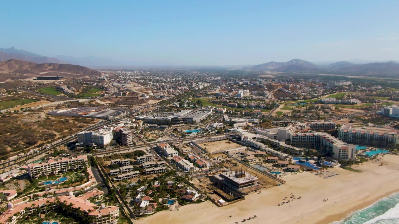 One second into the clip, the drone view holds over El Tule Beach Los Cabos with hotel blocks, blue pools, sandy shoreline, highway traffic, dry hills, and no visible people.