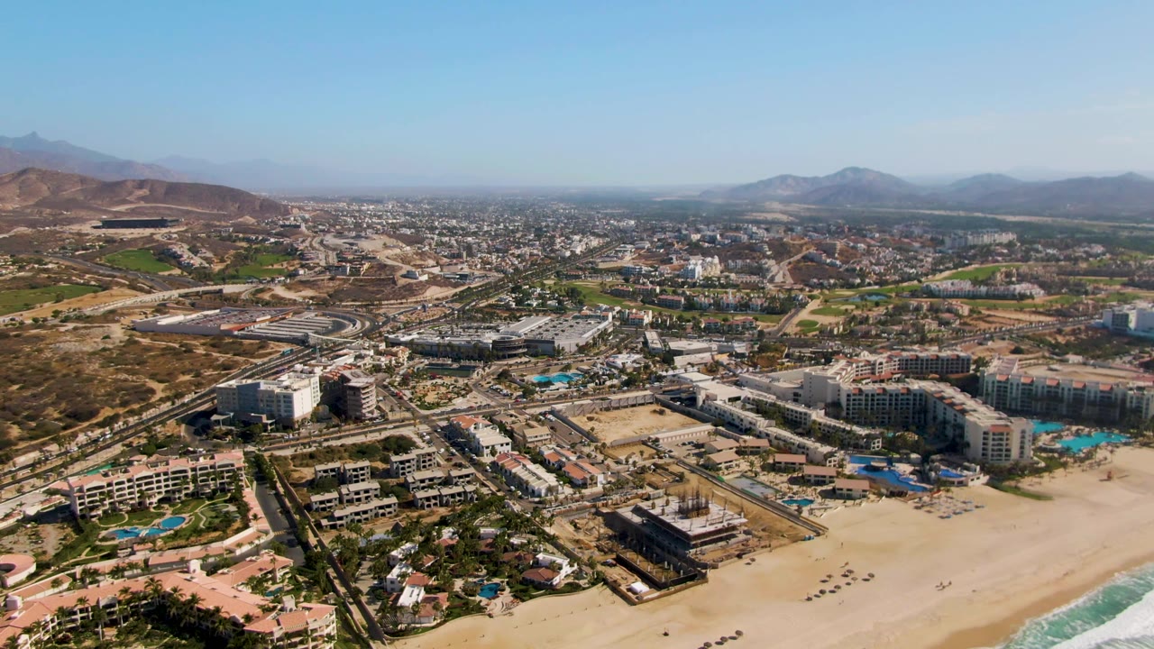 At 0:02, El Tule Beach Los Cabos appears from above with the beach and surf at lower right, resort pools and buildings across the middle, highway vehicles, and no visible people.