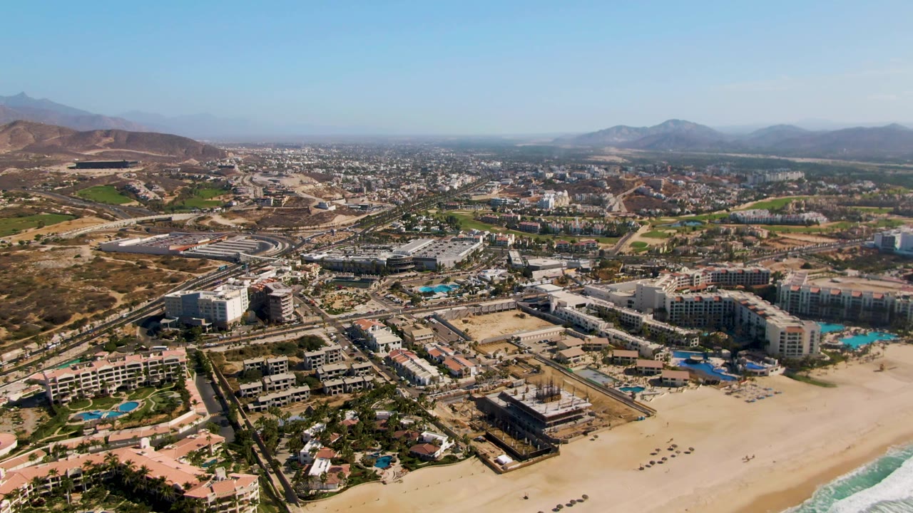 At 0:03, the aerial view of El Tule Beach Los Cabos shows more of the sandy beach foreground, with hotels, pools, a highway corridor, scattered vehicles, mountains, and no visible people.