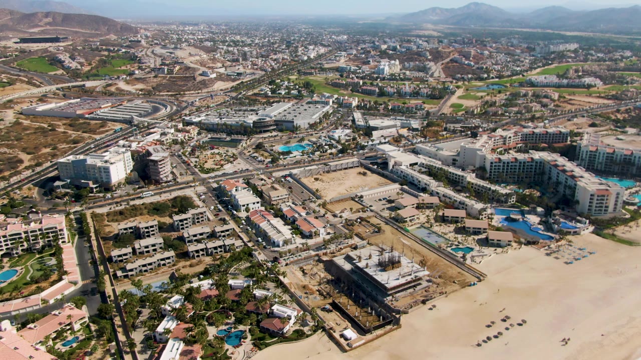 Aerial view of sandy beach, resort buildings, pools, roads, and hills.
