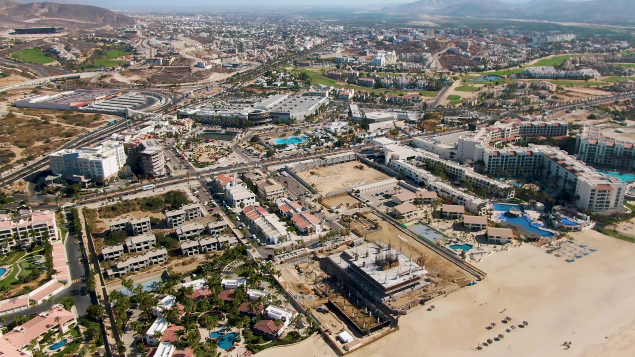 Aerial frame from El Tule Beach, Los Cabos, showing the beach foreground, several resort blocks with bright blue pools, inland streets, developed lots, distant hills, and no visible people.