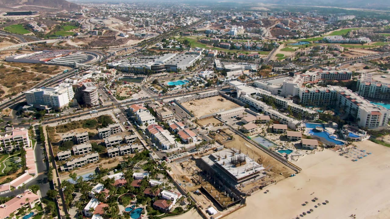 High drone perspective over El Tule Beach, Los Cabos, with a wide sandy foreground, resort complexes and pools across the center, roads and urban development behind them, and no visible people.