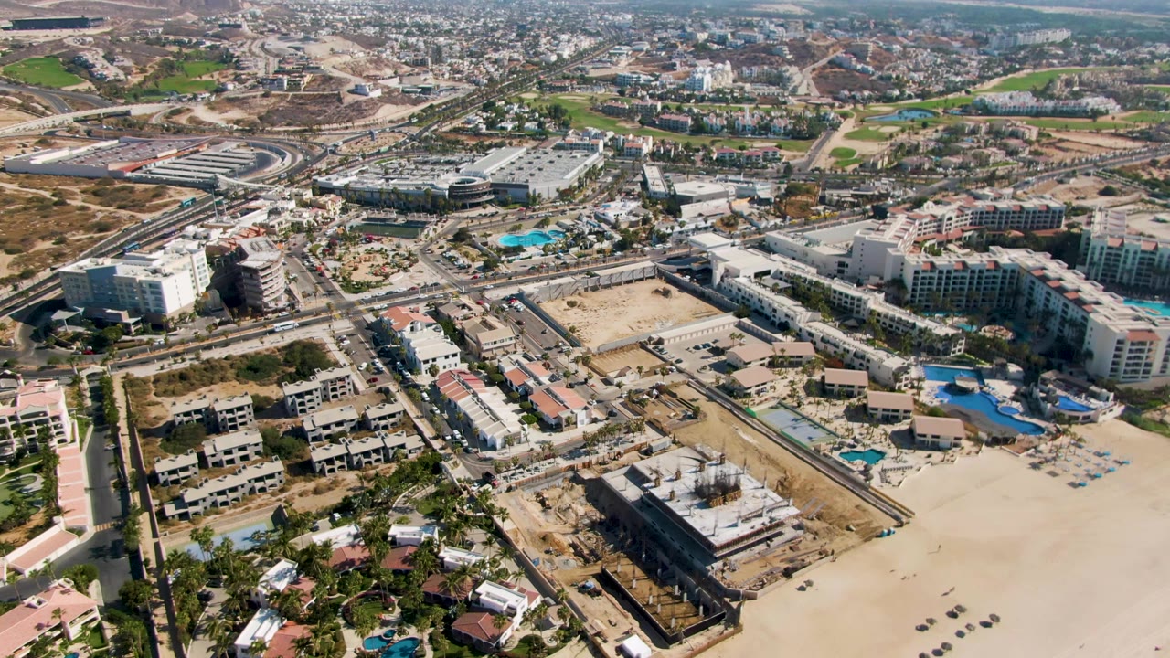 Mid-clip aerial over El Tule Beach, Los Cabos, showing the sandy shoreline area, hotel and resort structures, turquoise pools, road corridors, inland buildings, dry hills, and no visible people.