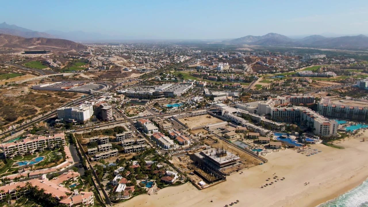 High aerial view over El Tule Beach Los Cabos with sandy beach in the foreground, resort pools and buildings inland, roads, dry hills, distant mountains, clear sky, and no visible people.