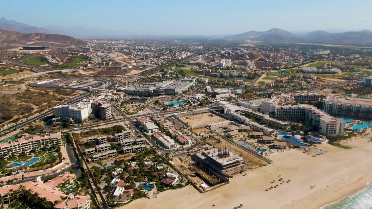 Aerial frame over El Tule Beach Los Cabos with the sandy shoreline low in frame, clustered resort buildings and blue pools near the coast, inland roads, mountains, clear sky, and no visible people.