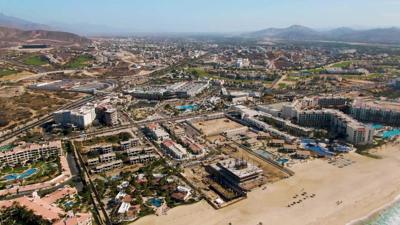 Wide drone view of El Tule Beach Los Cabos showing beach sand at the bottom, resort complexes and turquoise pools across the foreground, a road network, dry hills, distant mountains, and no visible people.