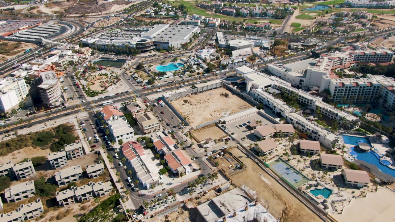 Opening aerial frame over El Tule Beach Los Cabos shows low-rise buildings, road intersections, turquoise pools, dry terrain, and distant hills under clear sky, with no visible people.