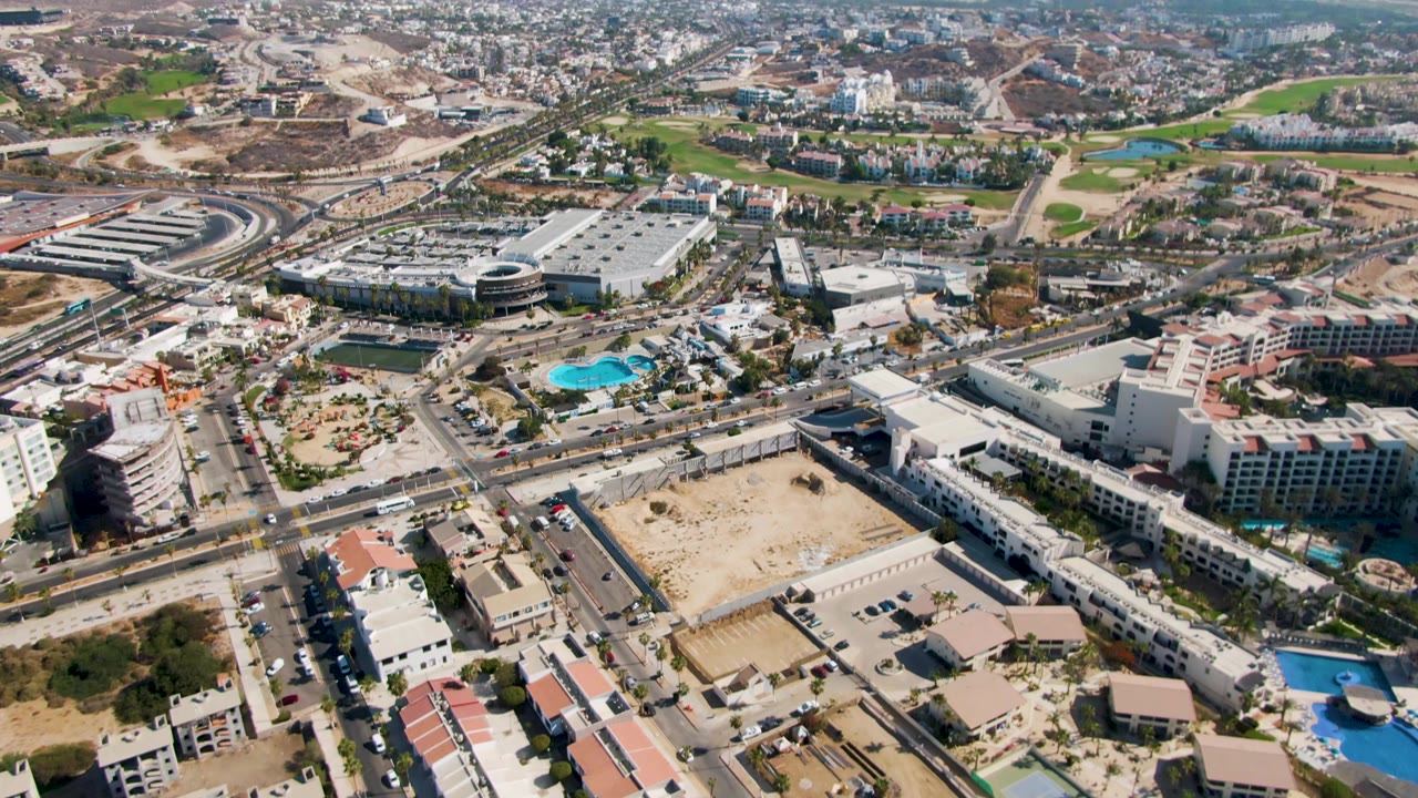 Third frame over El Tule Beach Los Cabos shows a larger sweep of the road network, hotel-like buildings, blue pools, open lots, greenery, and hazy hills, with no visible people.