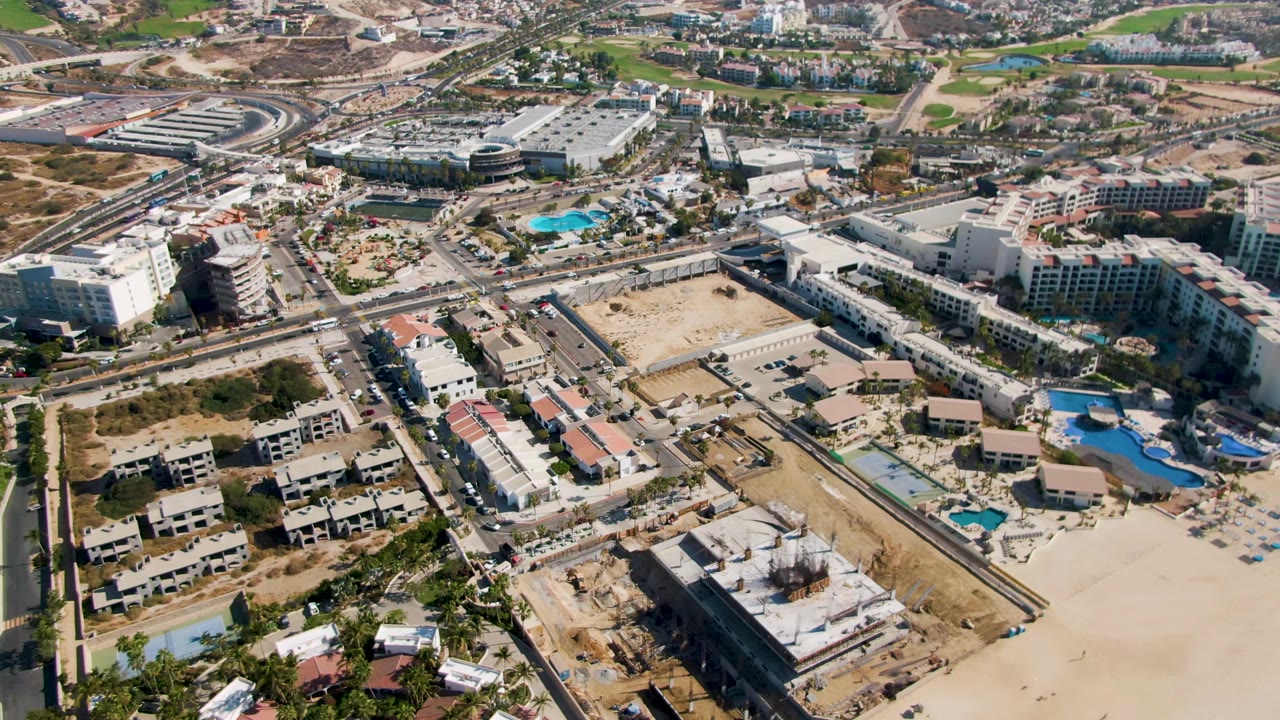 Aerial frame over El Tule Beach Los Cabos with a large sandy beach in the lower right, blue resort pools near center, roads crossing the scene, construction lots, and no visible people.