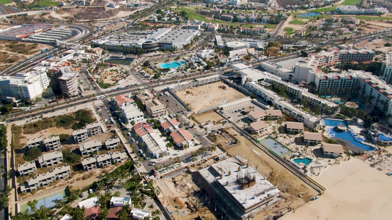 Oblique aerial view of El Tule Beach Los Cabos showing unfinished structures in the foreground, resort pools and buildings in the middle distance, roads with cars, sandy beach, and no visible people.