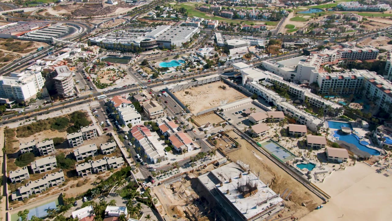 Mid-clip aerial view over El Tule Beach Los Cabos with construction sites at the bottom, a broad pale beach area to the right, resort pools near center, streets, parked or moving cars, and no visible people.