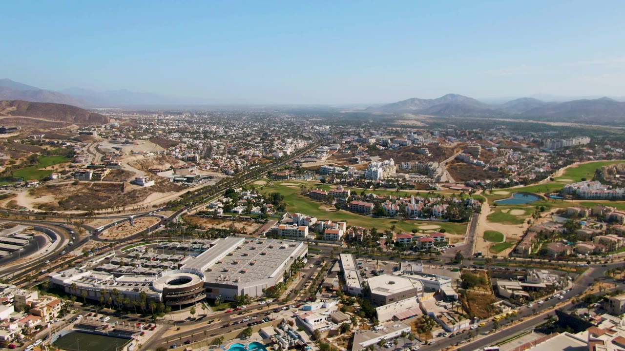 Opening aerial frame over El Tule Beach Los Cabos shows large commercial buildings, roads, golf course greens, dry hills, distant mountains, clear sky, and no visible people.