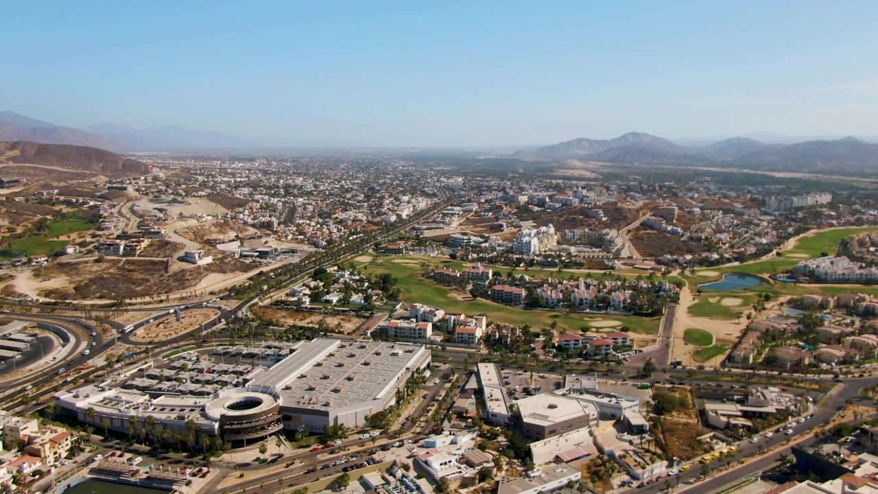 At 0:01, the El Tule Beach Los Cabos aerial view holds on roads and buildings in the foreground, golf course fairways and ponds in the middle distance, mountains on the horizon, and no visible people.