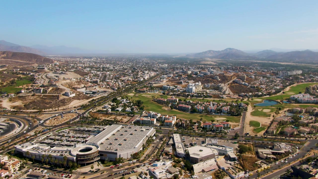 At 0:02, the high aerial frame shows El Tule Beach Los Cabos with a large roofed building at lower left, curving roads, golf greens and ponds, hillside construction areas, distant mountains, and no visible people.