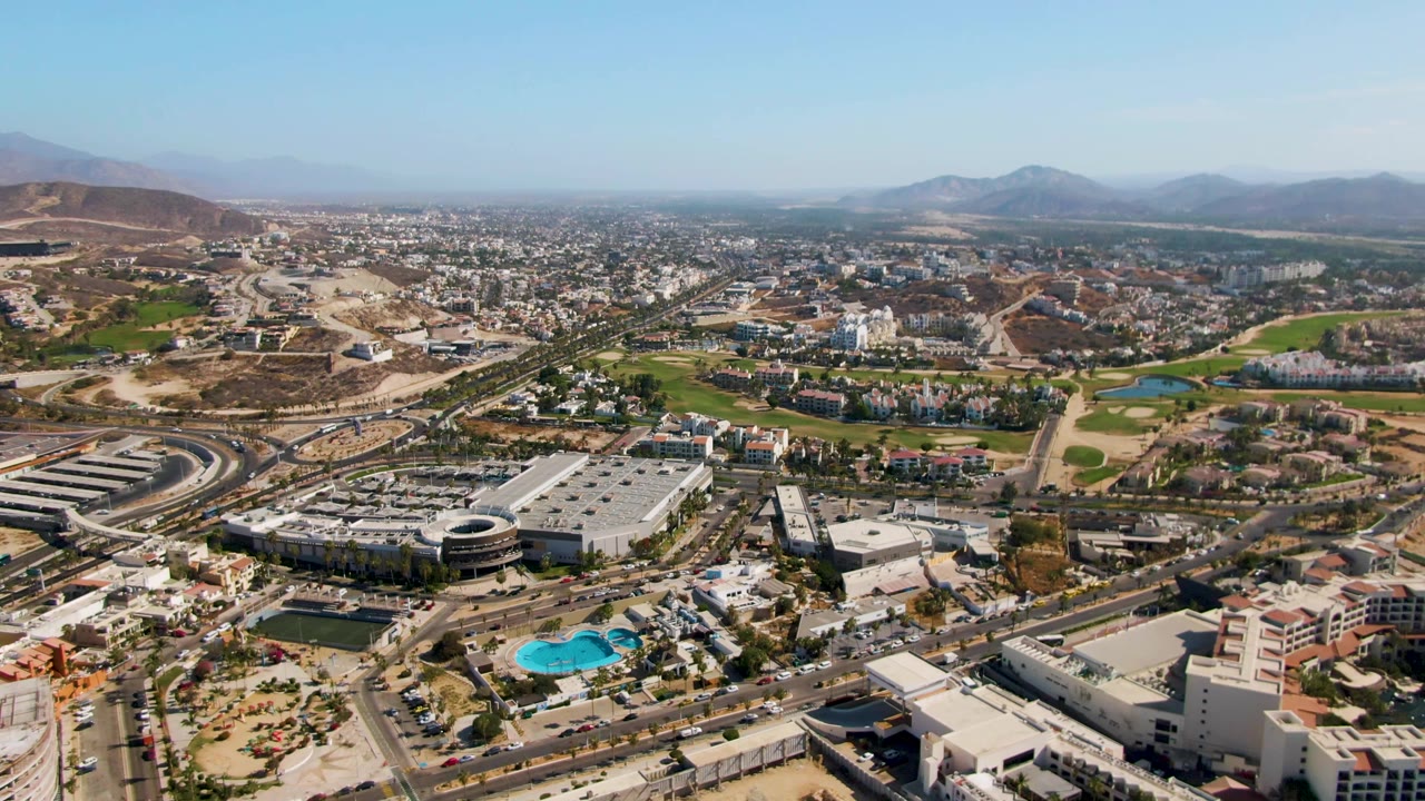 High aerial view over El Tule Beach Los Cabos with roads, large buildings, blue pools, green landscaped areas, dry hills, distant mountains, clear sky, and no visible people.