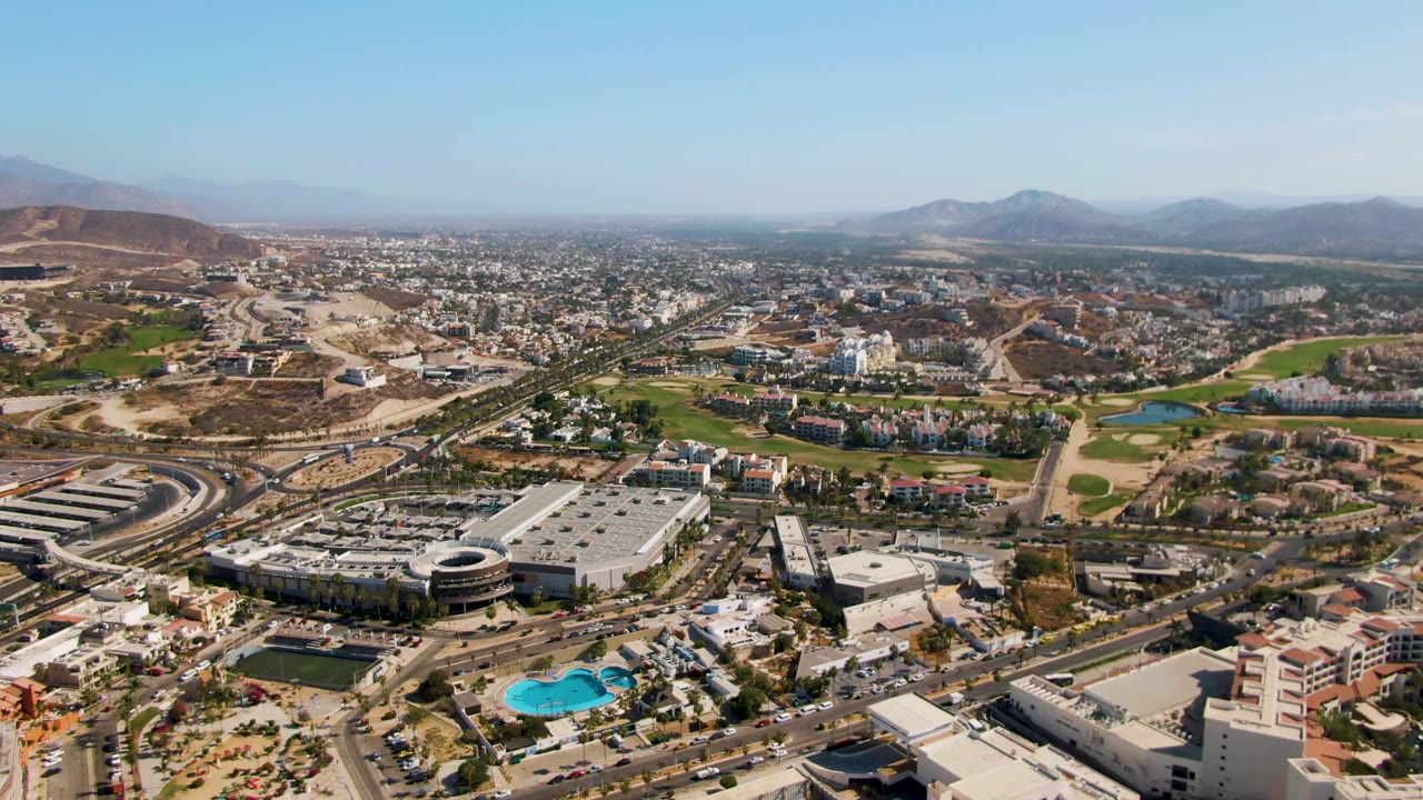 Wide aerial frame over El Tule Beach Los Cabos showing dense city blocks, road corridors, green lawns, a small blue pond or pool area, dry slopes, mountains, clear sky, and no visible people.