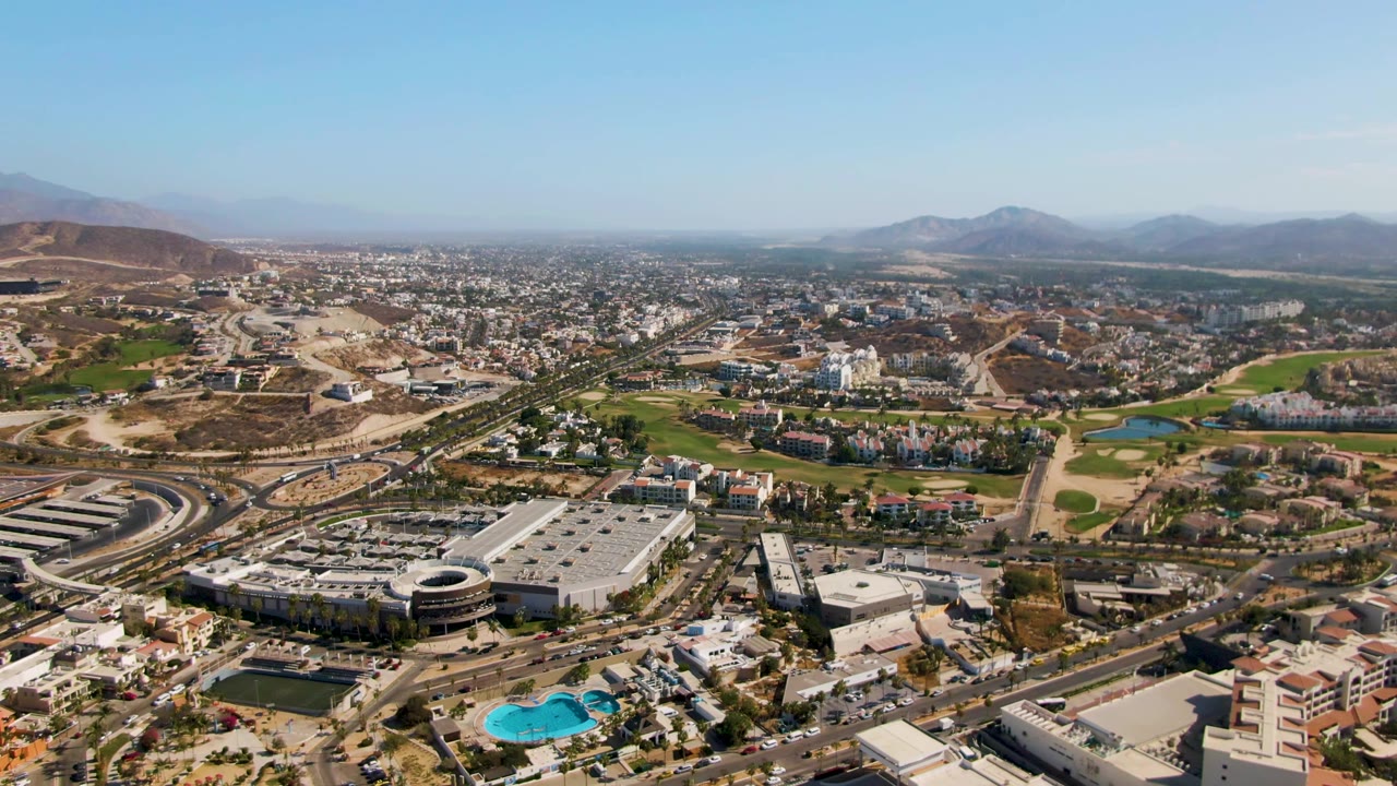 Mid-clip aerial view of El Tule Beach Los Cabos with a large light-roofed building in the foreground, curving roads, residential areas, turquoise pools, mountains, clear sky, and no visible people.