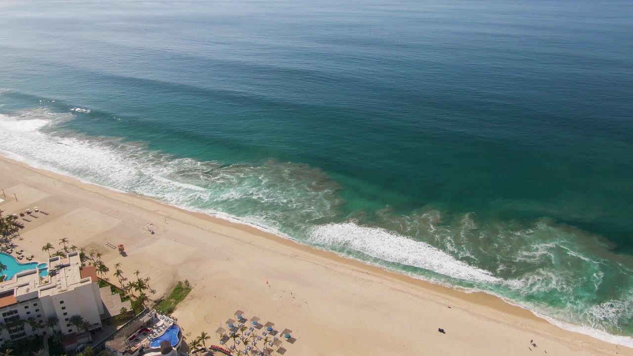 At 0:00, an aerial view of El Tule Beach in Los Cabos shows deep blue ocean, white surf, pale sand, resort buildings, pools, palm trees, umbrellas, and no visible people.