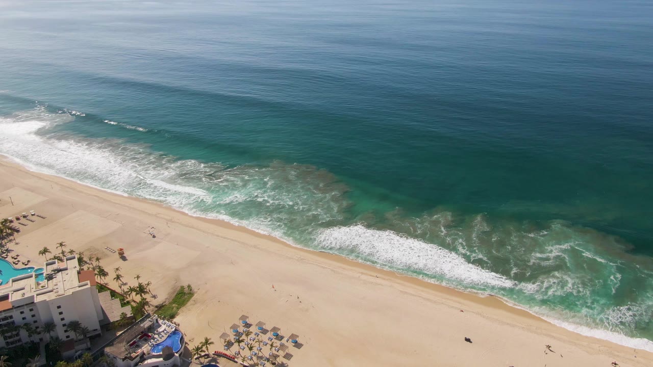At 0:01, the El Tule Beach shoreline shows a longer white foam line across the sand, with blue ocean above, resort roofs and umbrellas at the lower left, and no visible people.