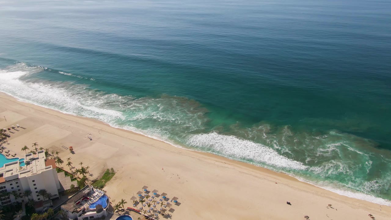 At 0:02, foamy water curls onto the sandy shore at El Tule Beach, with open blue water above, resort buildings and pools at the bottom left, and no visible people.