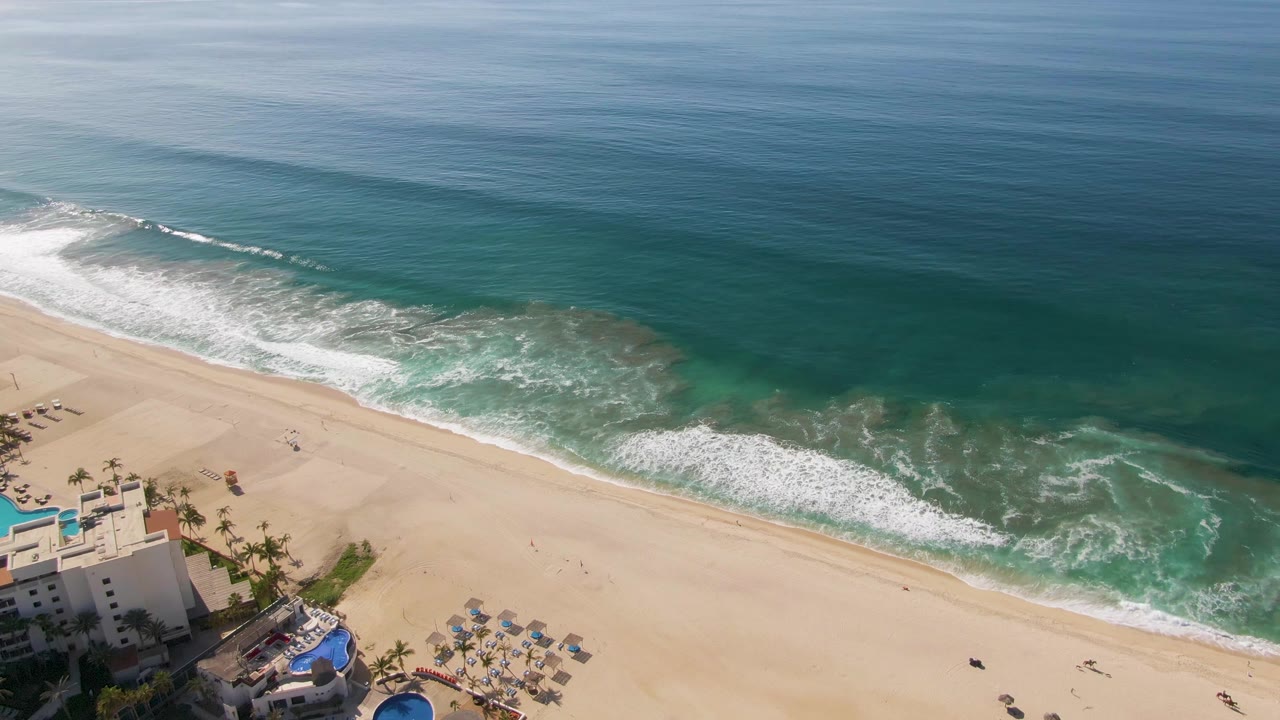 Aerial beach frame with breaking waves and resort buildings along the lower left edge.