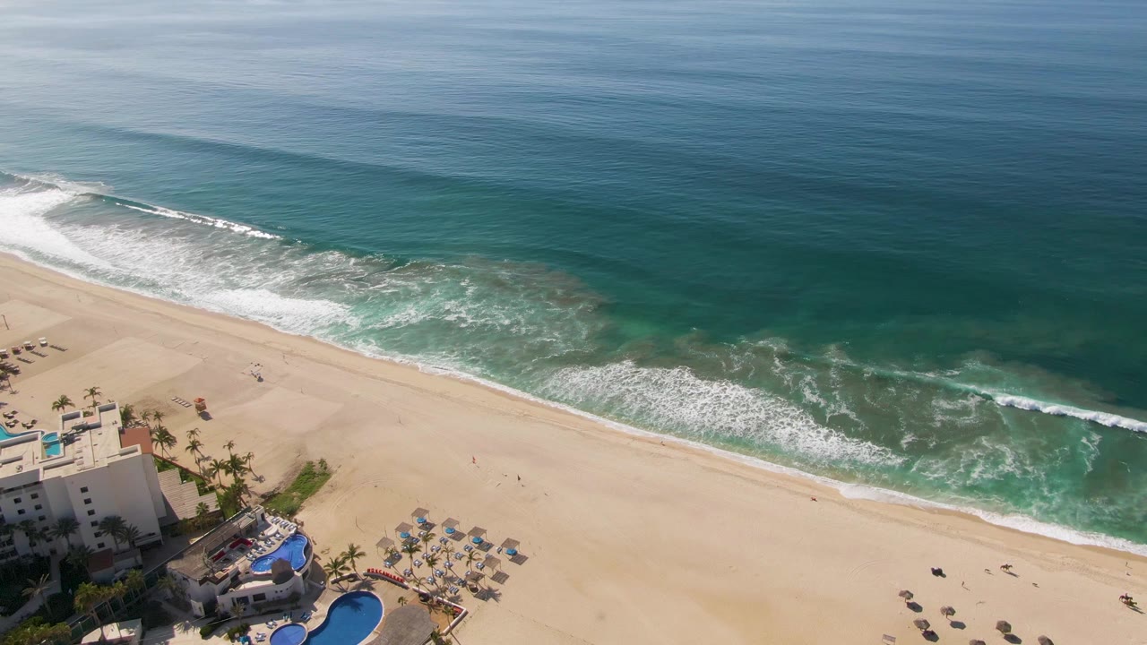 Aerial view of sandy beach, surf, ocean, and a partial resort pool area.