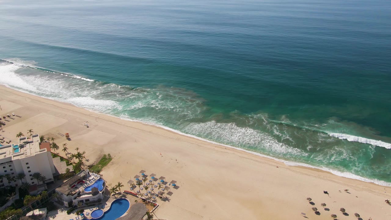 Aerial view of El Tule Beach in Los Cabos with the resort pool and umbrellas still visible at lower left, white surf crossing the sand, and no visible people.