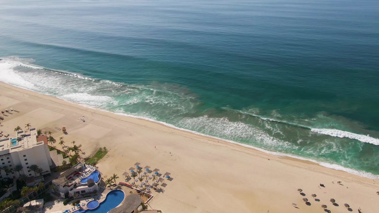 El Tule Beach appears from above with a wide band of sand, foamy waves, deeper blue water offshore, and the resort corner lower left with no visible people.