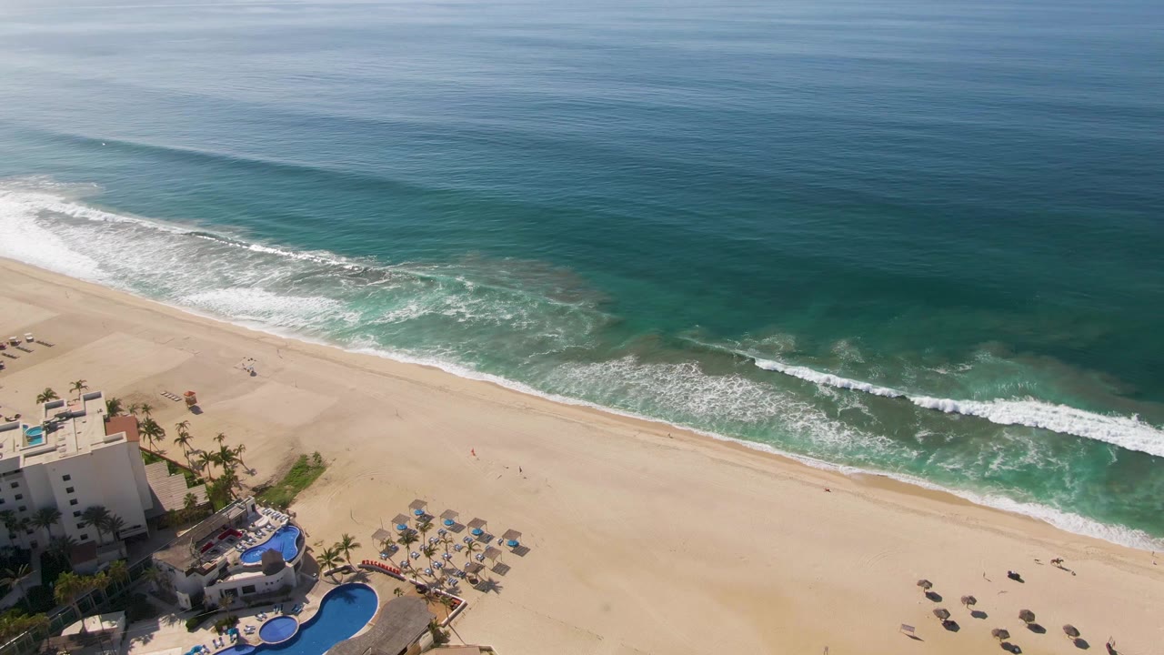 A later aerial frame over El Tule Beach shows more open ocean above the diagonal shoreline, with the resort pool partly cropped at lower left and no visible people.