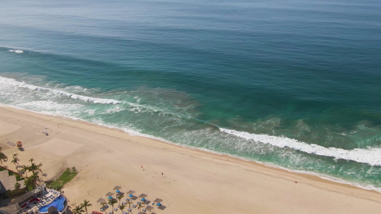 Aerial frame over El Tule Beach in Los Cabos showing blue-green ocean, a diagonal band of white surf, pale sand, small beachgoers, umbrellas, palms, and a pool edge at the lower left.
