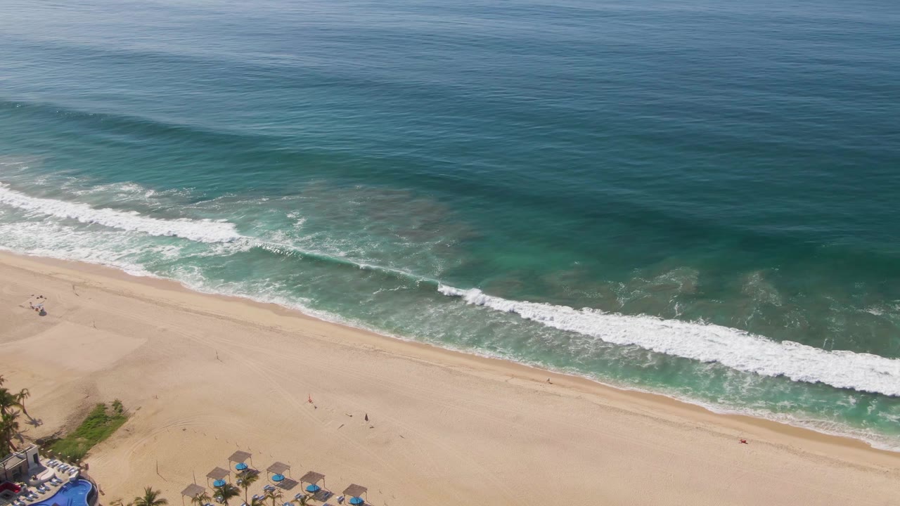 Aerial frame from El Tule Beach in Los Cabos with darker ocean at the top, translucent green shallows near the break, a clean diagonal foam line, and a few tiny people on the sand.