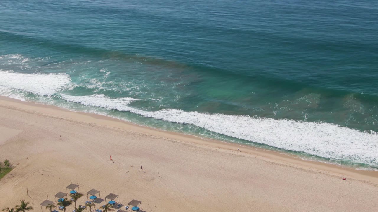 Aerial frame from El Tule Beach in Los Cabos showing blue-green ocean, white surf, tan sand, and a small section of resort umbrellas at the bottom edge.