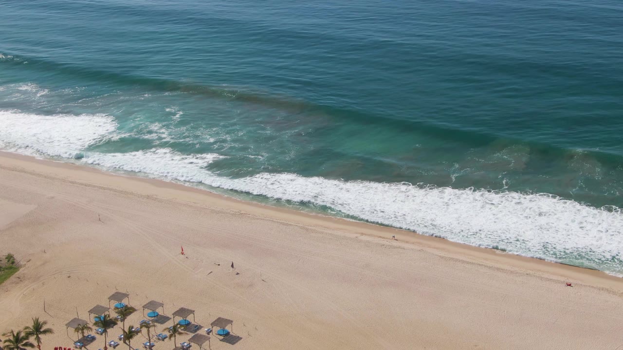 Foamy waves roll toward a broad beach with umbrellas and palm trees beginning to show below.