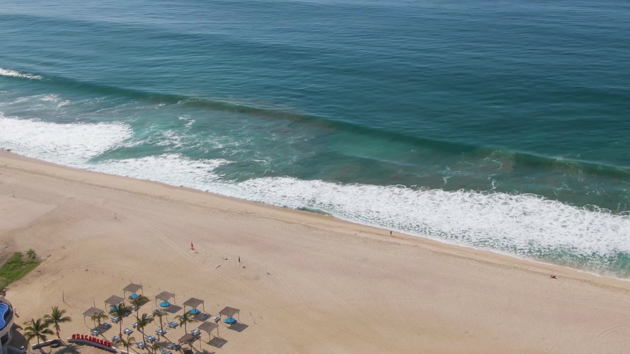 El Tule Beach appears from above with rolling surf, pale sand, scattered tiny beachgoers, a cluster of umbrellas, palm trees, and the curved edge of a resort pool at lower left.