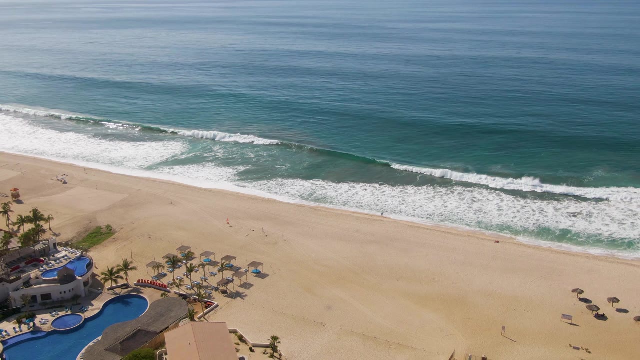 Aerial frame over El Tule Beach in Los Cabos showing resort pools, palms, umbrellas, open sand, and white surf bands, with no visible people.