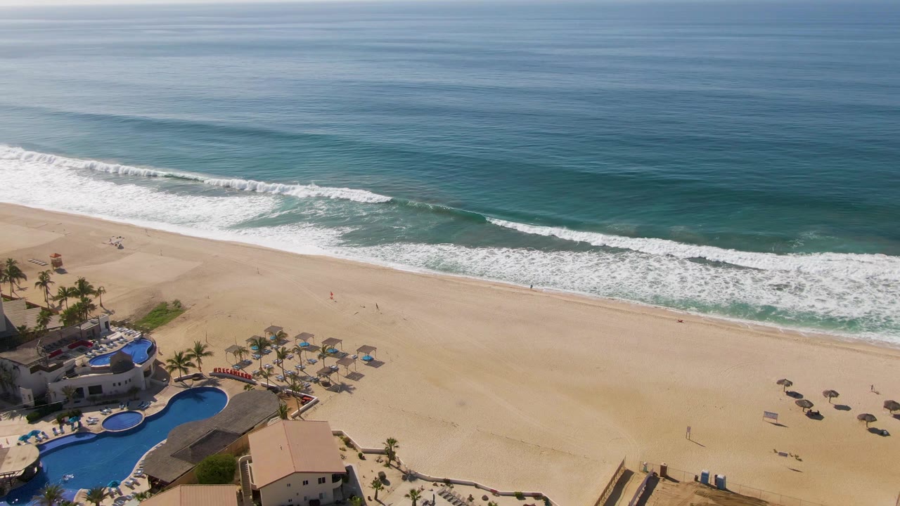 Aerial view at El Tule Beach with the blue pool complex lower left, umbrellas on the sand, and a broad line of breaking waves offshore, with no visible people.