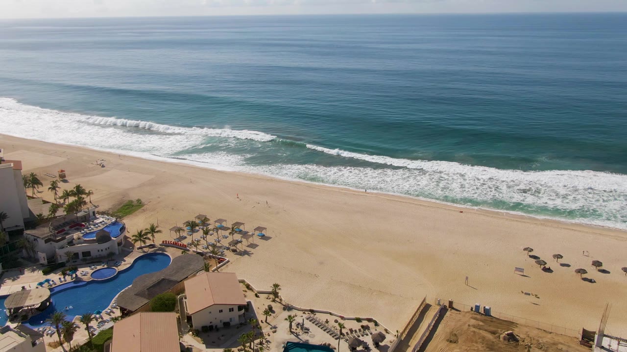 Elevated Los Cabos beach frame with resort rooftops and pools at the bottom, umbrellas scattered across the sand, and smooth blue ocean beyond the surf, with no visible people.