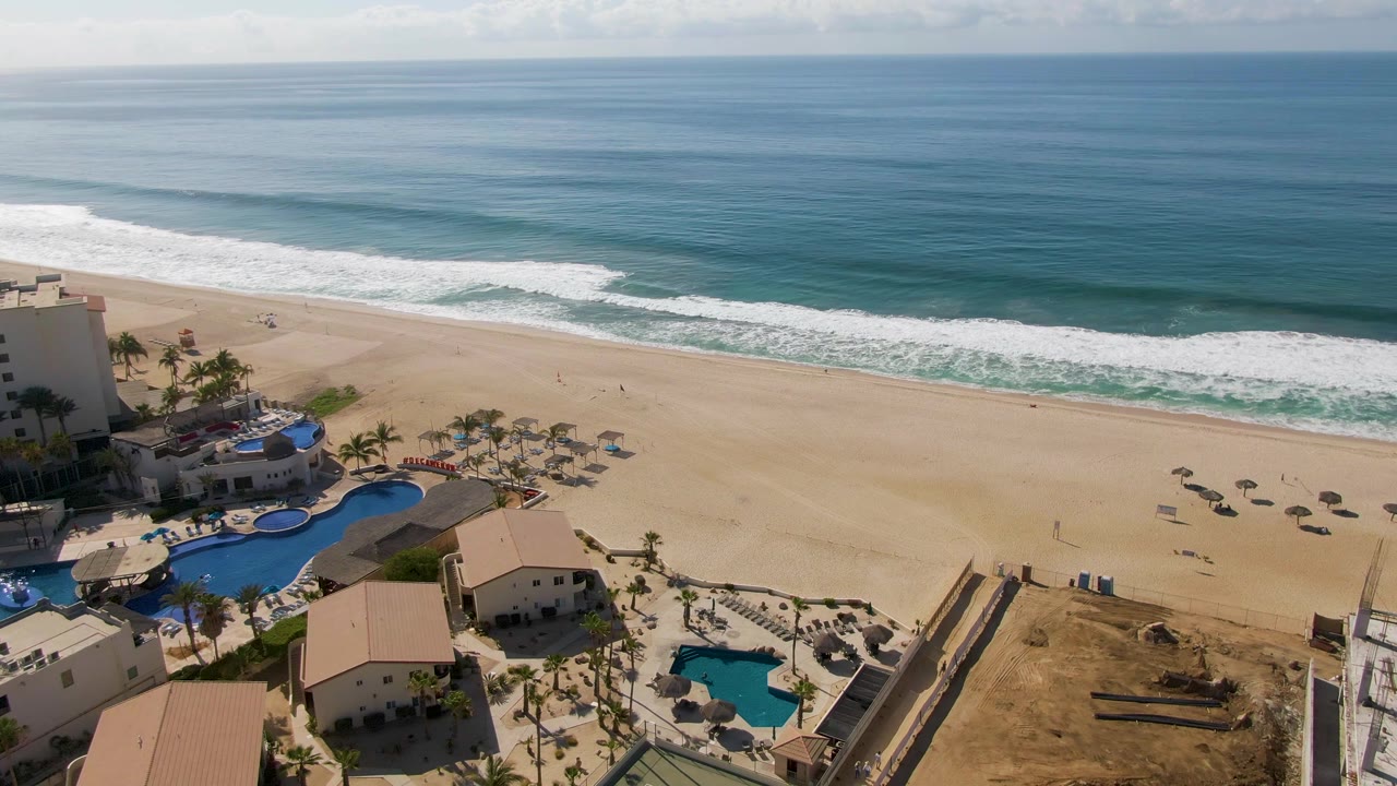 Aerial view over El Tule Beach in Los Cabos with blue ocean, white surf, tan sand, scattered umbrellas, tiny beachgoers, palm trees, pools, and light-colored buildings at the start of the clip.