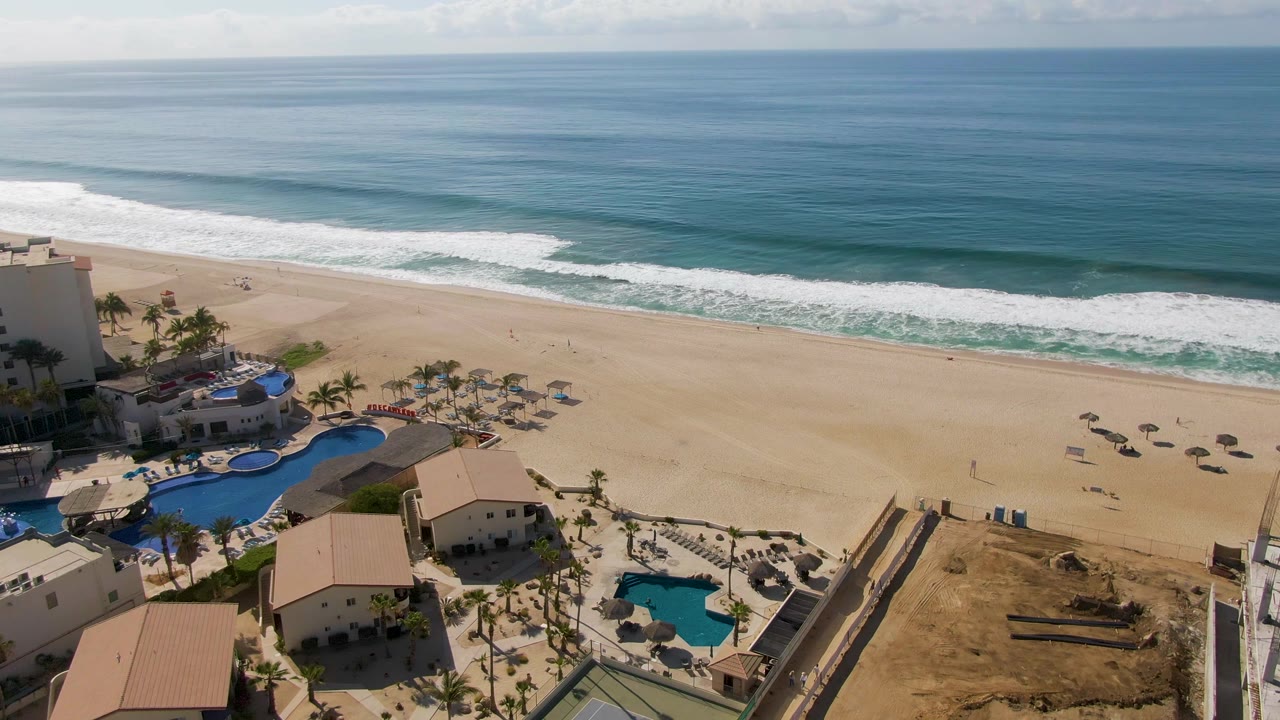 Wide aerial beach view with calm ocean, breaking waves, umbrellas, pools, and palms.