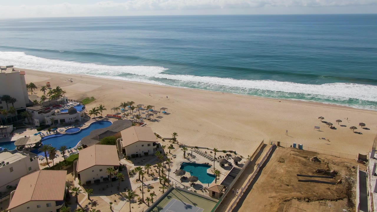Drone-style aerial view of El Tule Beach in Los Cabos with the ocean horizon high in frame, a foamy wave line across the shore, scattered umbrellas on sand, and pool areas beside pale buildings.