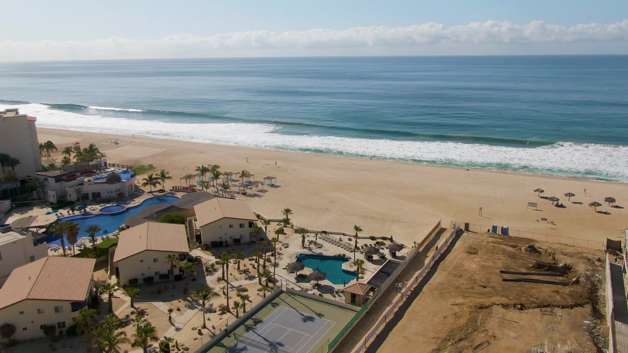 Aerial frame over El Tule Beach in Los Cabos with resort buildings, pools, palm trees, tennis court, sandy beach, blue ocean, white surf, and no visible people.
