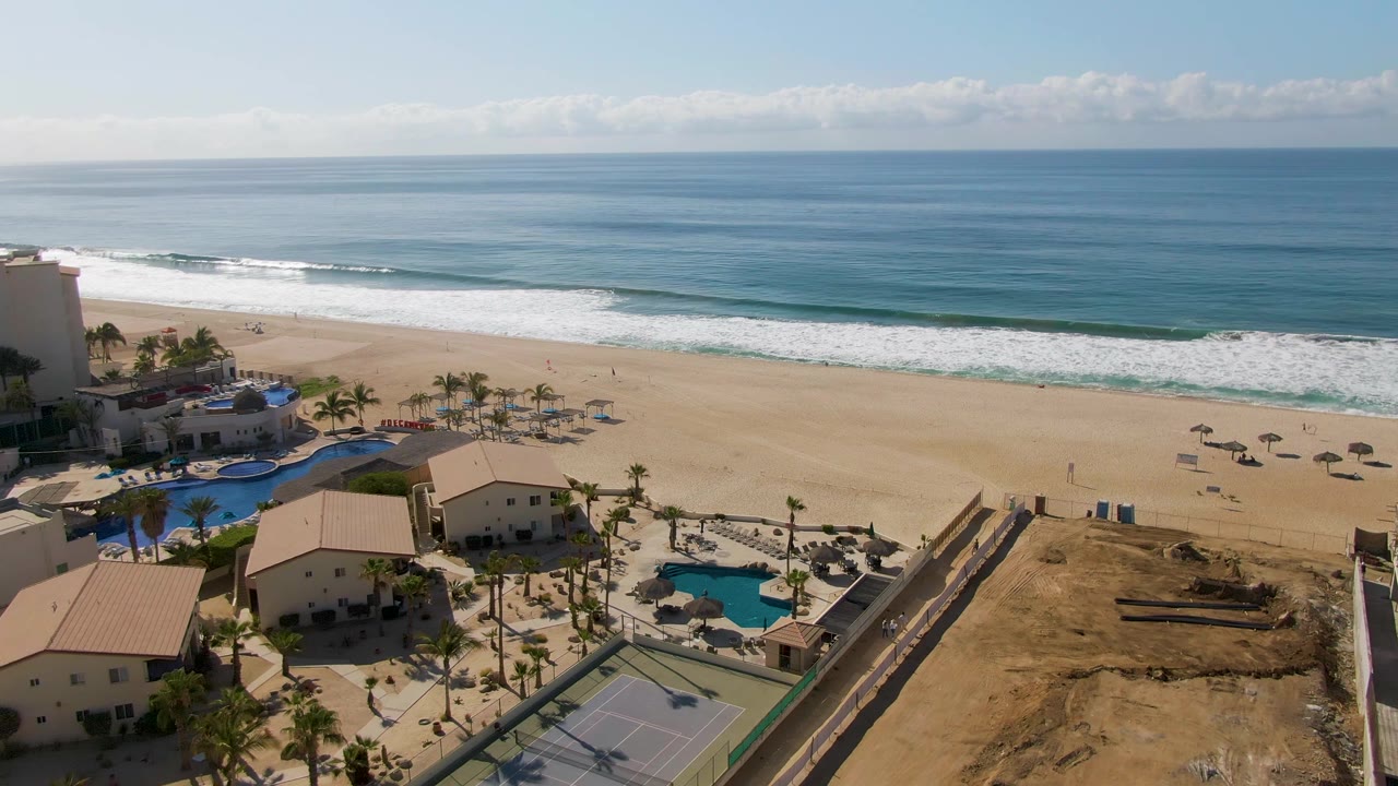 A slightly later aerial view of El Tule Beach shows whitecaps moving toward the sand, with resort pools, palms, umbrellas, a tennis court, and no visible people.